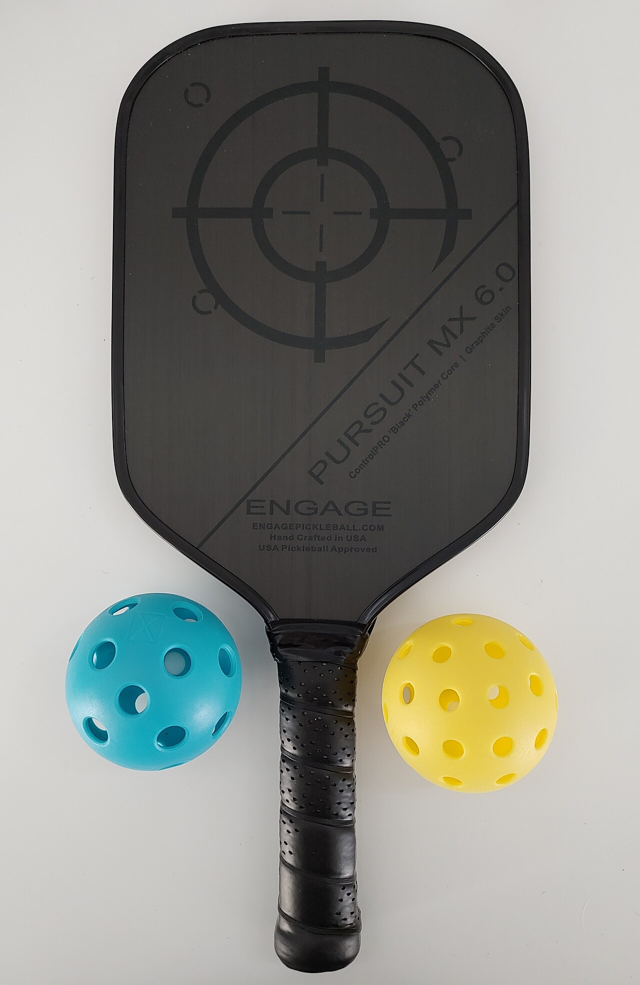 A Pursuit MX 6.0 pickleball paddle by Engage with one blue indoor pickleball and one yellow outdoor pickleball. The blue ball has 26 round holes, and 